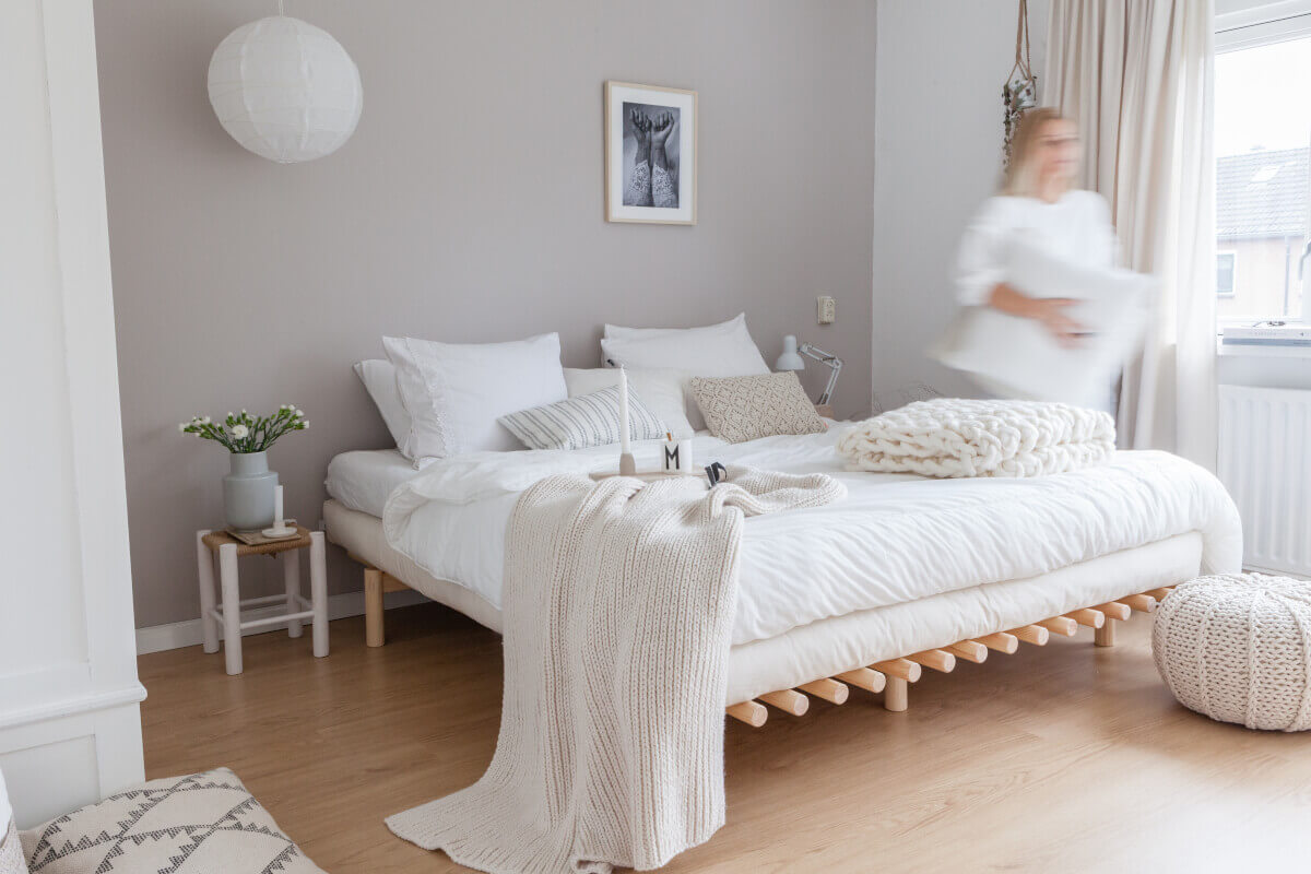 Wnętrze sypialniane w estetyce scandi chic: proste, drewniane łóżko w naturalnym kolorze, funkcjonalność, białe ściany, jasna podłoga i pastele, które szykownie przełamują całość.